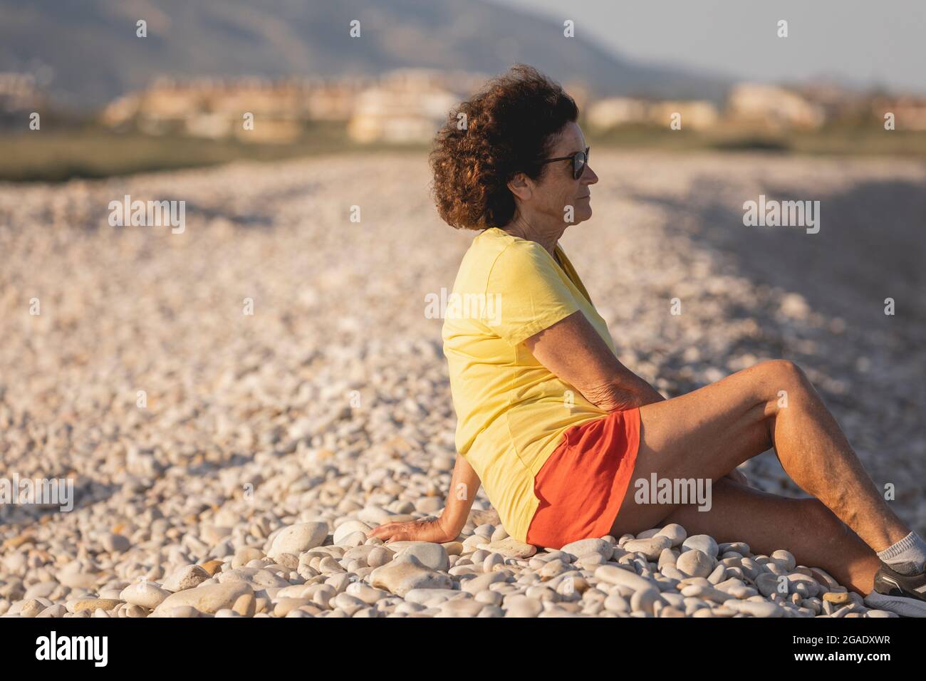 Una donna allegra e vitale, nei suoi anni '70, è seduta su una spiaggia di ghiaia naturale al tramonto, guardando fuori al Mar Mediterraneo, Spagna Foto Stock