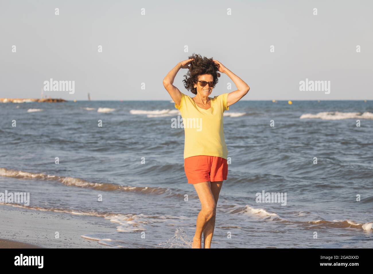 Una donna allegra e vitale, nei suoi anni '70, cammina su una spiaggia al tramonto, sulle rive del Mar Mediterraneo, Spagna Foto Stock