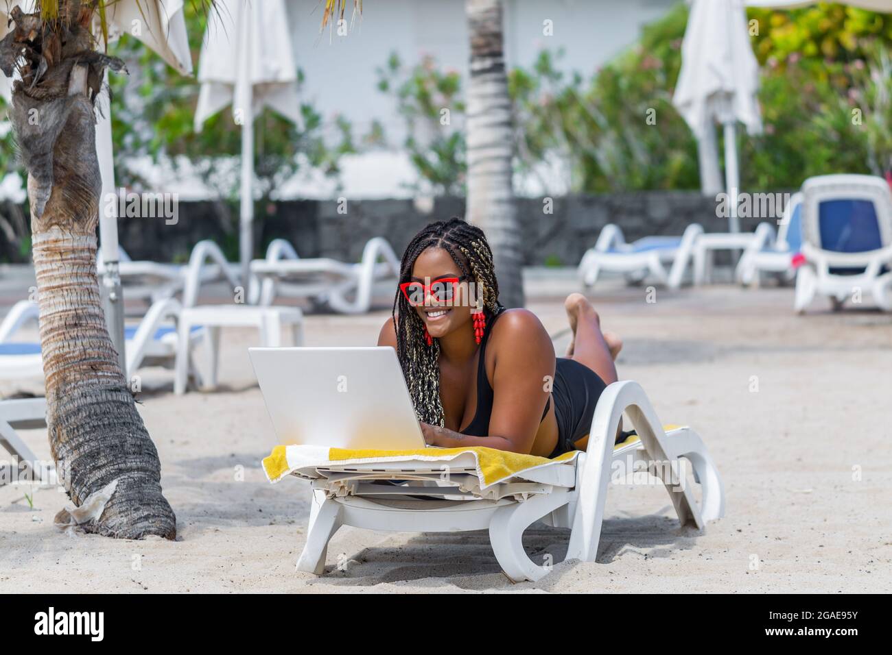 Donna afroamericana sulla spiaggia sdraiata su una sedia a sdraio utilizzando un computer portatile. Donna adulta allegra che si gode una giornata estiva in spiaggia. Foto Stock