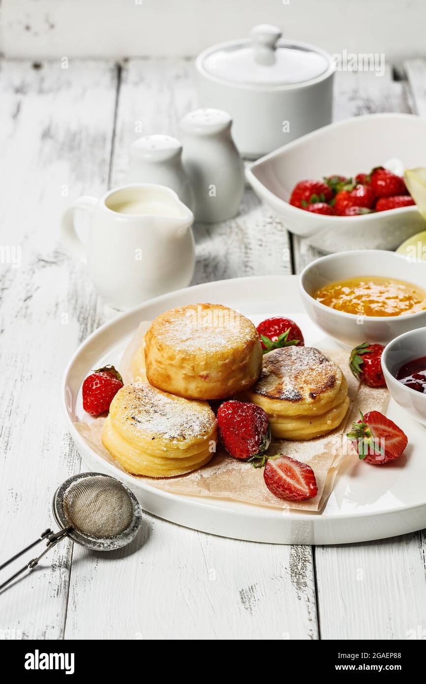Frittelle di formaggio cottage con fragole fresche e marmellata. Frittelle di formaggio di cagliata su un piatto bianco cosparso di zucchero in polvere. Deliziosa pausa fatta in casa Foto Stock