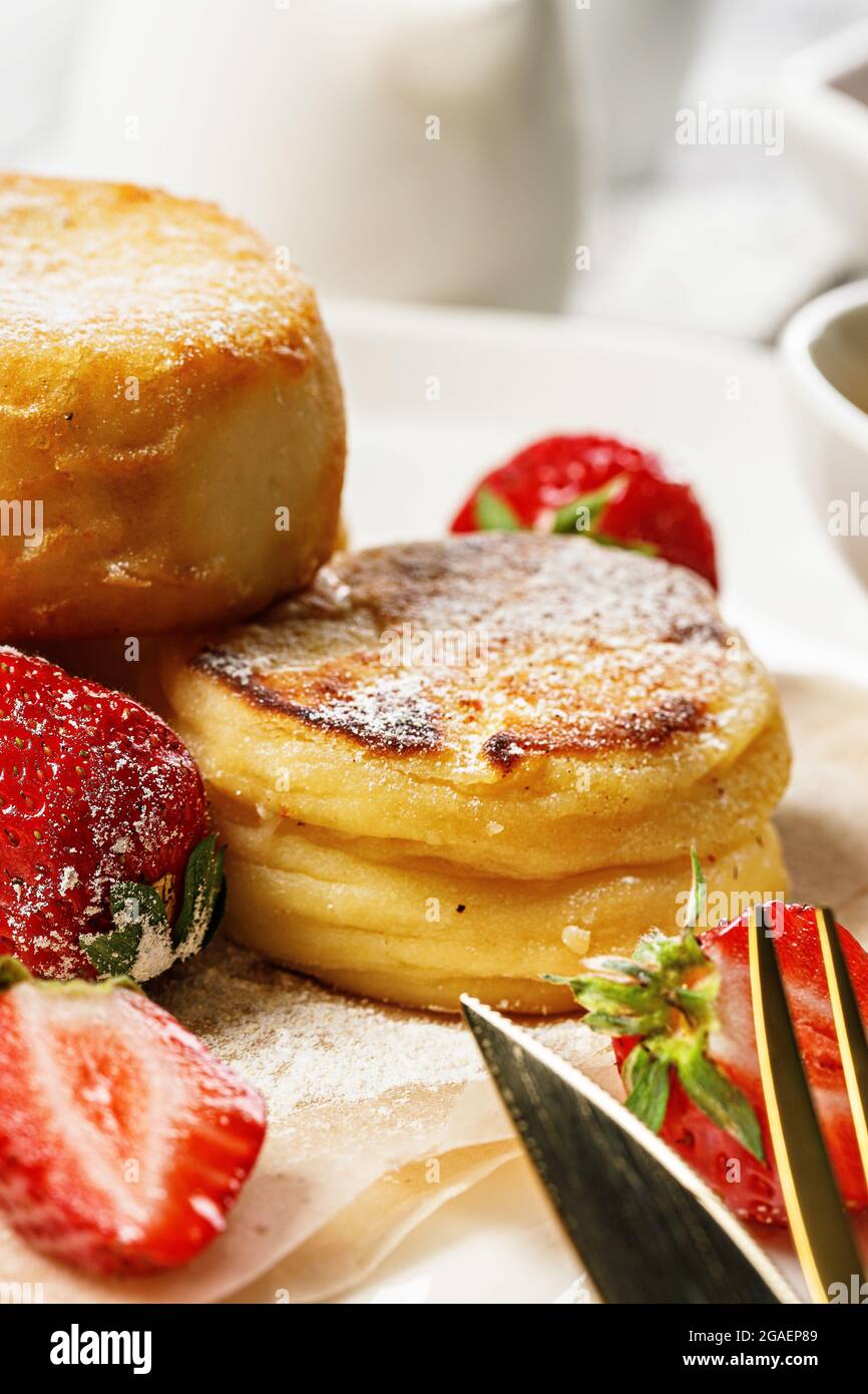 Primo piano frittelle di formaggio Cottage con fragole fresche e marmellata. Frittelle di formaggio di cagliata su un piatto bianco cosparso di zucchero in polvere. Deliziosa casa Foto Stock