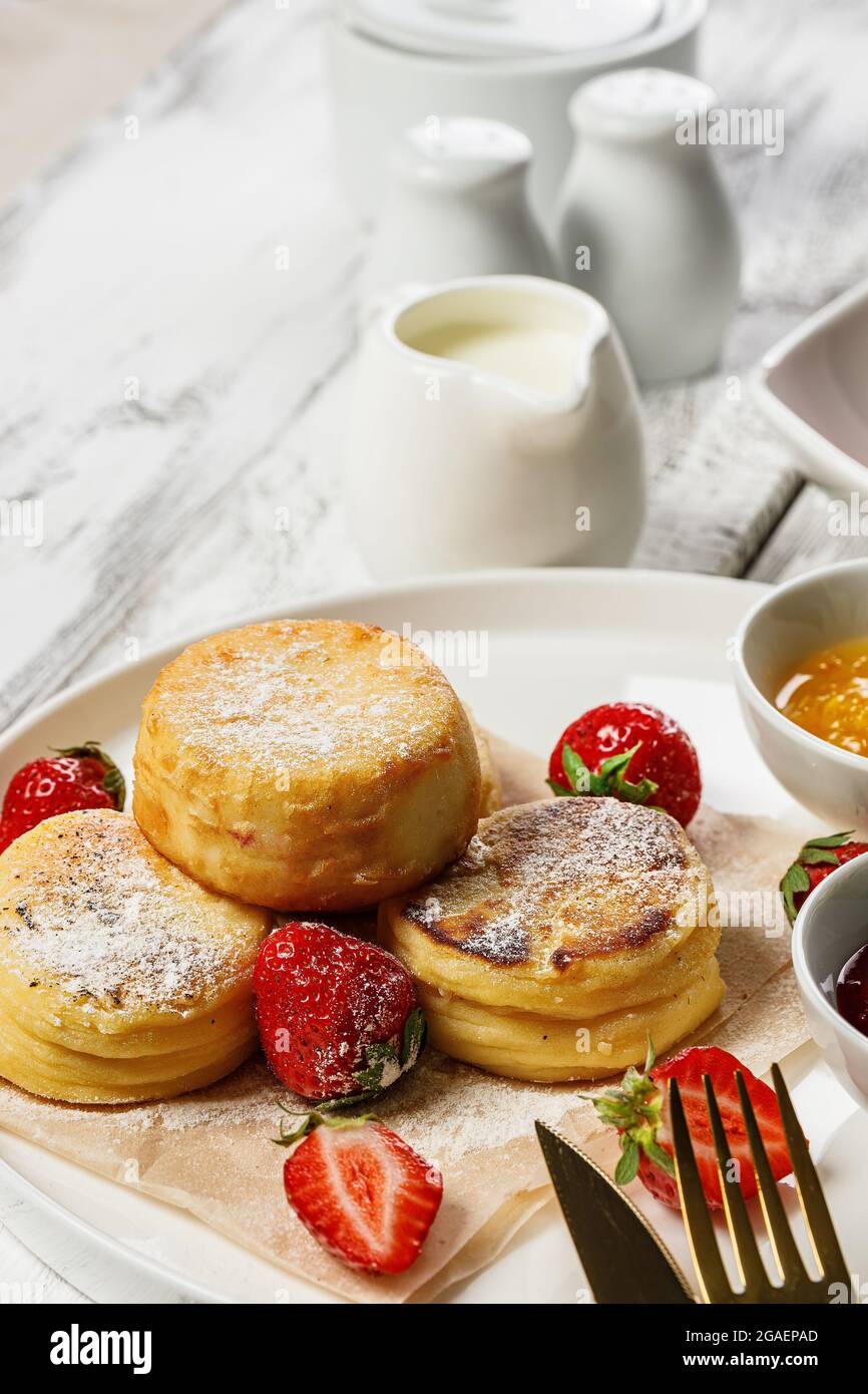 Frittelle di formaggio a scatto verticale con fragole fresche e marmellata. Frittelle di formaggio di cagliata su un piatto bianco cosparso di zucchero in polvere. Delizioso Foto Stock