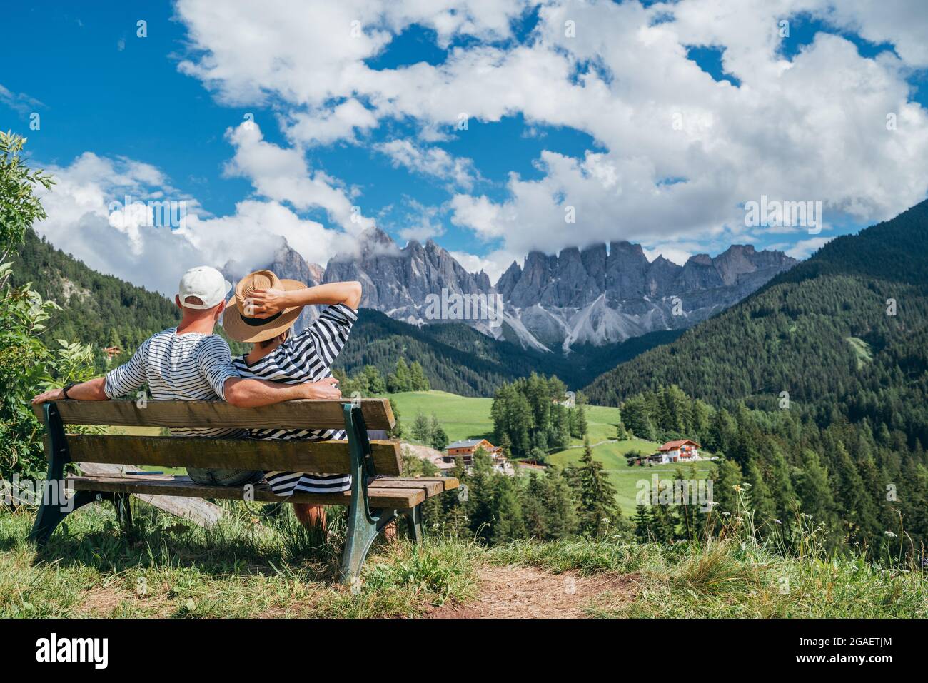 Coppia avvolgente con cime bianche a righe vestite seduta sulla panchina godendo la vista del villaggio di Santa Maddalena e le splendide e pittoresche Alpi dolomitiche in Val di Fune Foto Stock