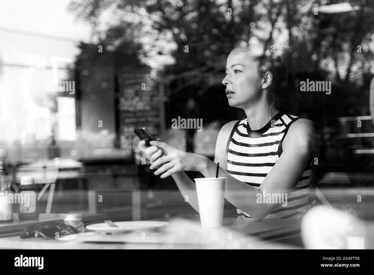 Premurosa donna caucasica tenendo il telefono cellulare mentre si cerca attraverso la caffetteria finestra durante la pausa caffè. Foto Stock