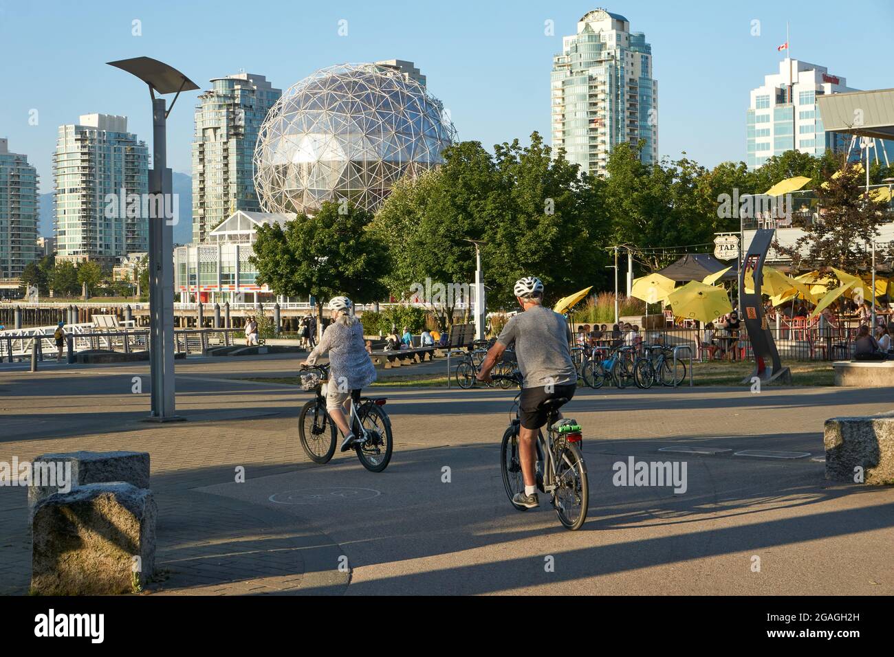 Uomo anziano e donna che cavalcano le loro biciclette nel Villaggio di False Creek, Vancouver, British Columbia, Canada Foto Stock