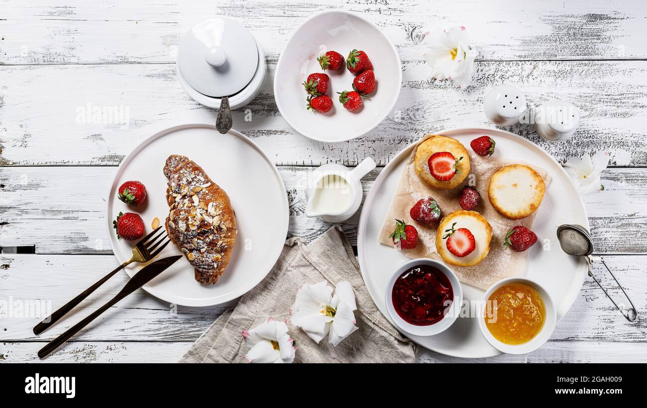 Banner alimentare. Appetitosi croissant croccanti e frittelle al formaggio di Cottage con fragole fresche e marmellata di frutti di bosco. Colazione deliziosa e sana. Int Foto Stock