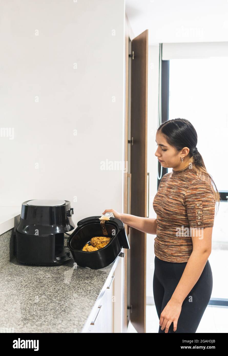 Giovane donna adolescente che tira il pollo fritto con i cereali fuori dalla friggitrice ad aria Foto Stock