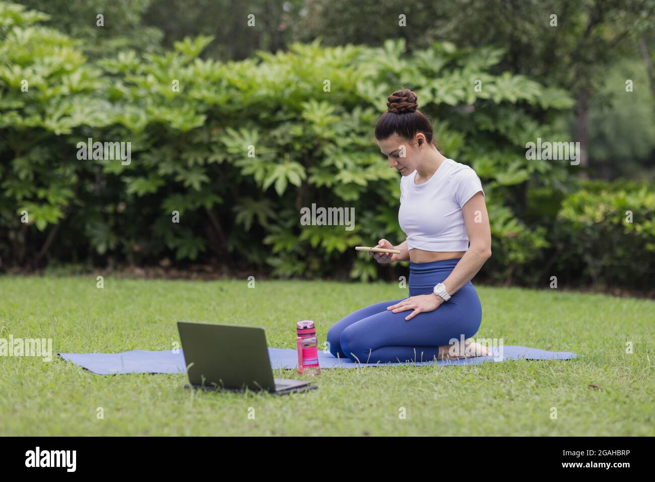 Giovane donna caucasica che si esercita online nel parco estivo durante la pandemia di coronavirus COVID-19. Social distancing durante l'allenamento fitness. Ragazza che indossa Foto Stock