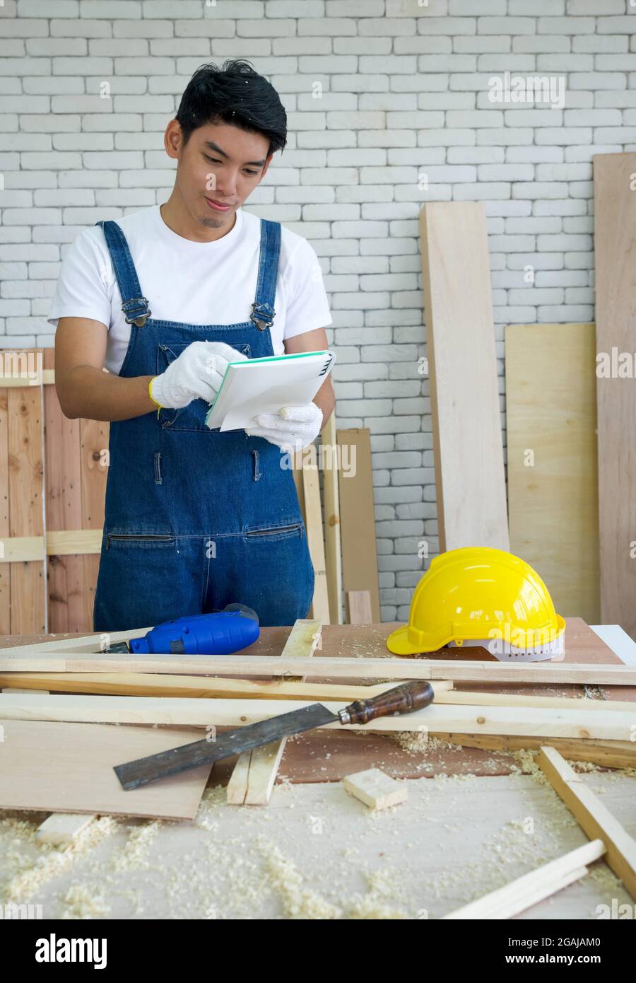 Il falegname asiatico ispeziona l'elenco degli articoli che devono essere fatti come programmato, consegna il lavoro con un volto felice. Atmosfera di lavoro mattutina in officina Foto Stock