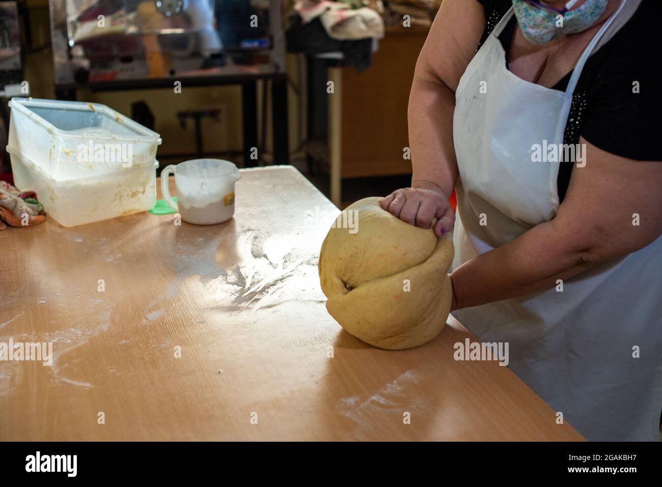 Mujer caucásica masando masa para hacer pan y bizcochos en una mesa color madera con luz de día, en una panadería. Foto Stock