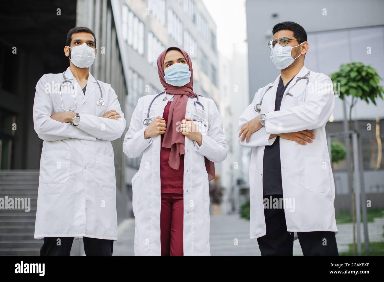 Vista frontale di tre operatori sanitari multietnici sicuri in abiti e scrub medici, indossando maschere per il viso, in posa con braccia incrociate all'aperto di fronte al moderno edificio ospedaliero. Covid-19 pandemia Foto Stock