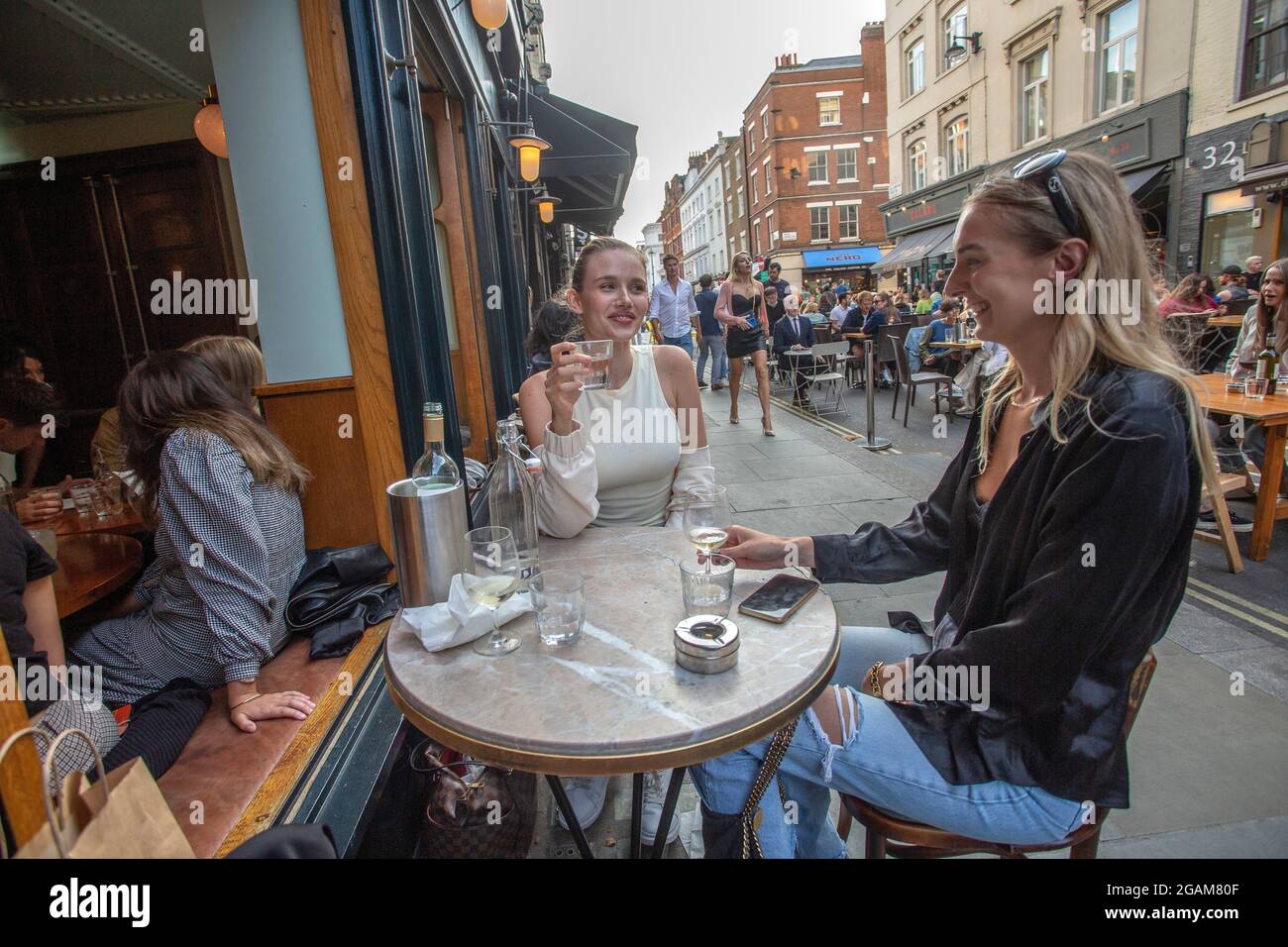 Due giovani donne che celebrano il 'giorno della libertà' che termina oltre un anno di restrizioni COVID-19 in Inghilterra persone che bevono sui tavoli posti fuori o Foto Stock