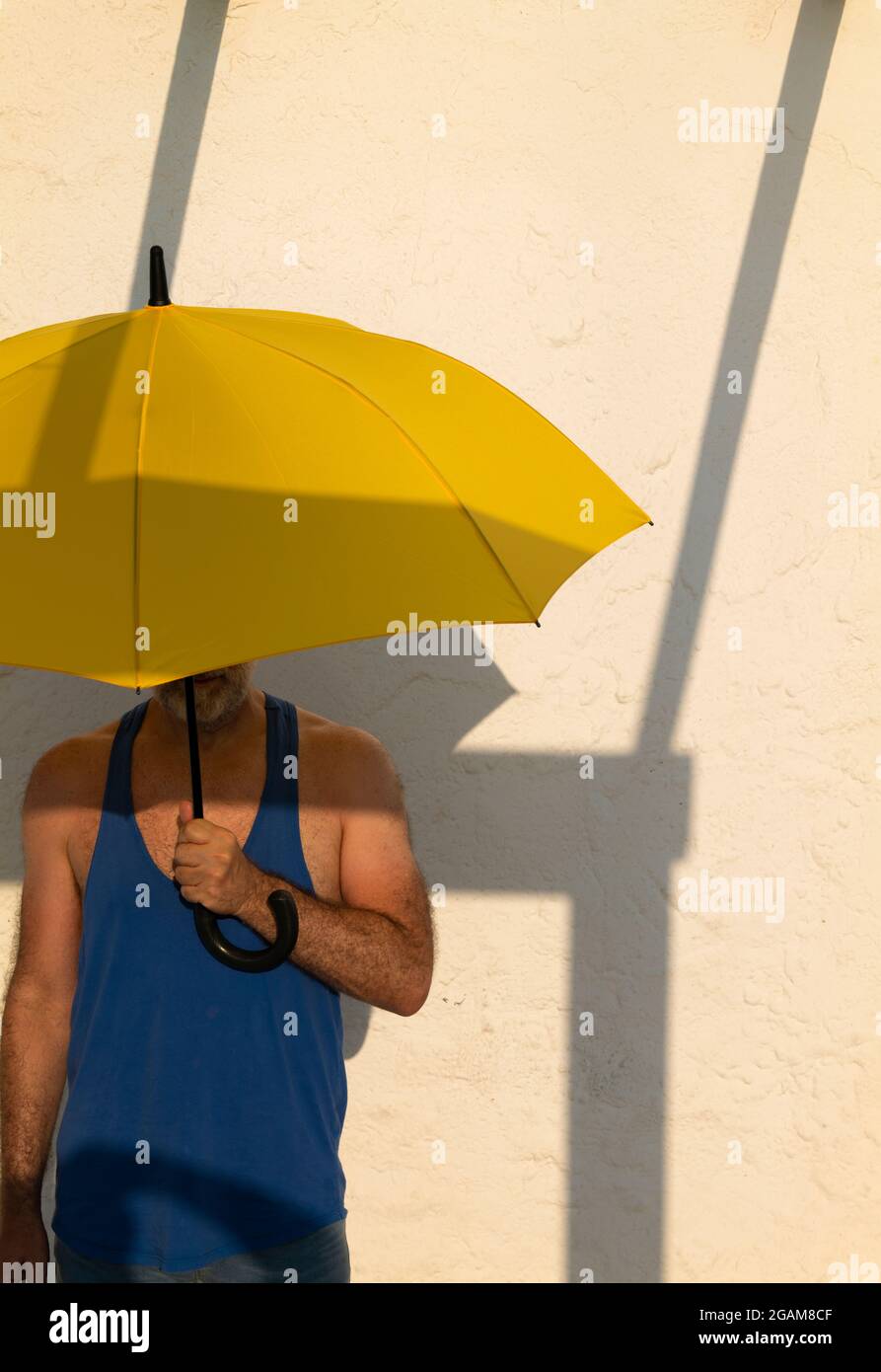 Uomo con canotta blu che tiene un ombrello giallo davanti a una parete bianca con ombra Foto Stock