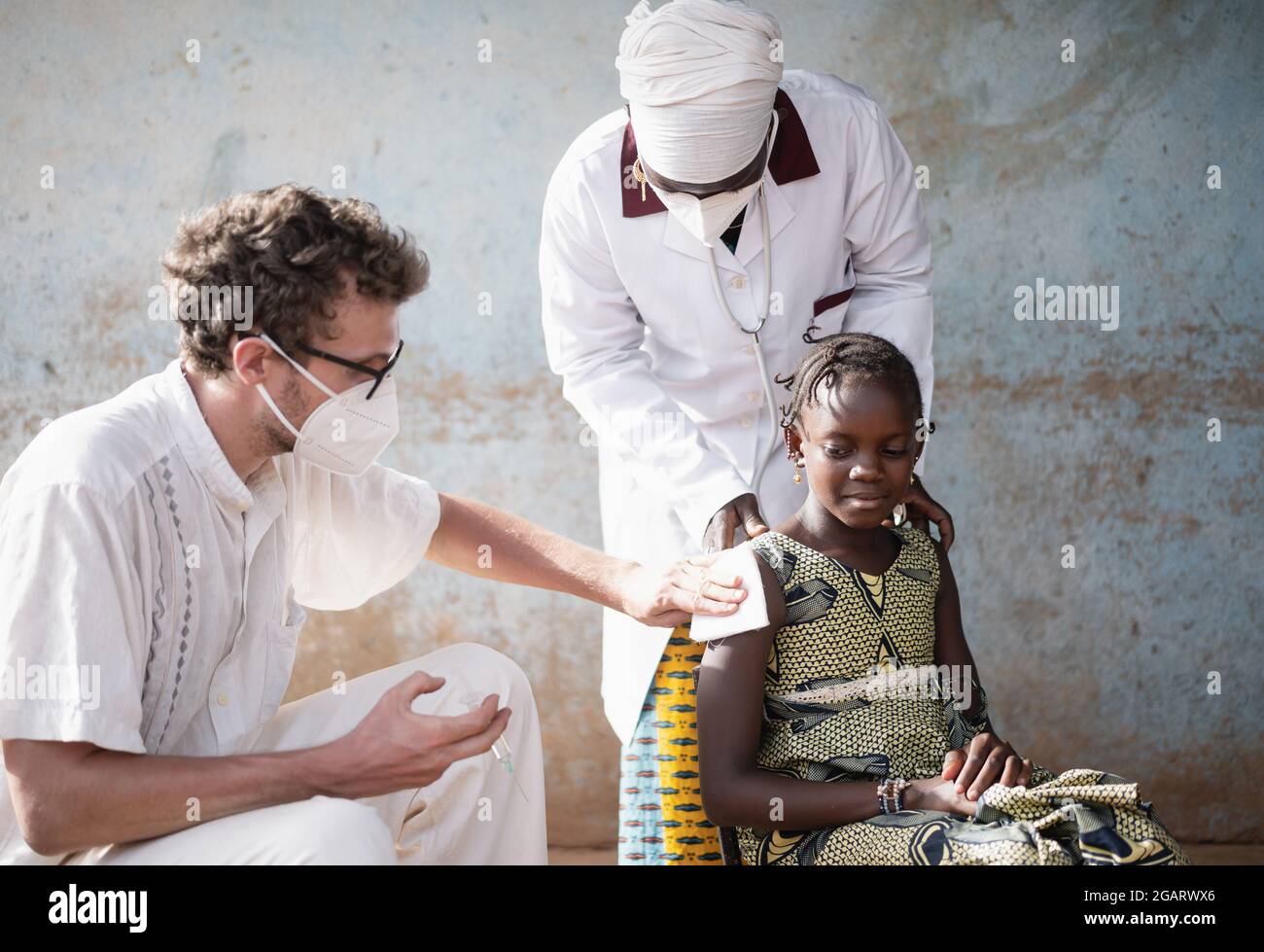 In questa immagine, un giovane aiuto umanitario bianco con maschera facciale sta insegnando metodi di disinfezione e iniezione ad un infermiere africano sul braccio di una com Foto Stock