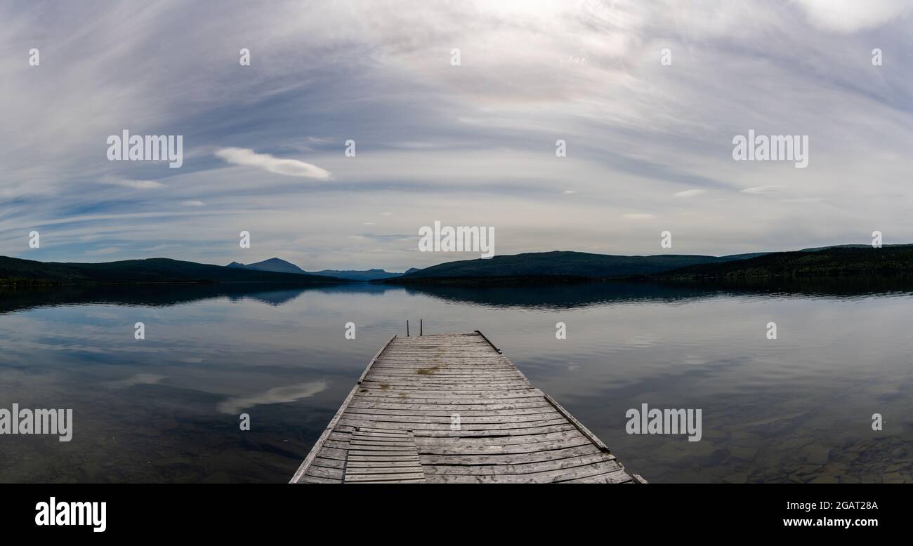 Un paesaggio panoramico di un lago tranquillo con riflessi di montagne e cielo e un molo in legno in primo piano Foto Stock