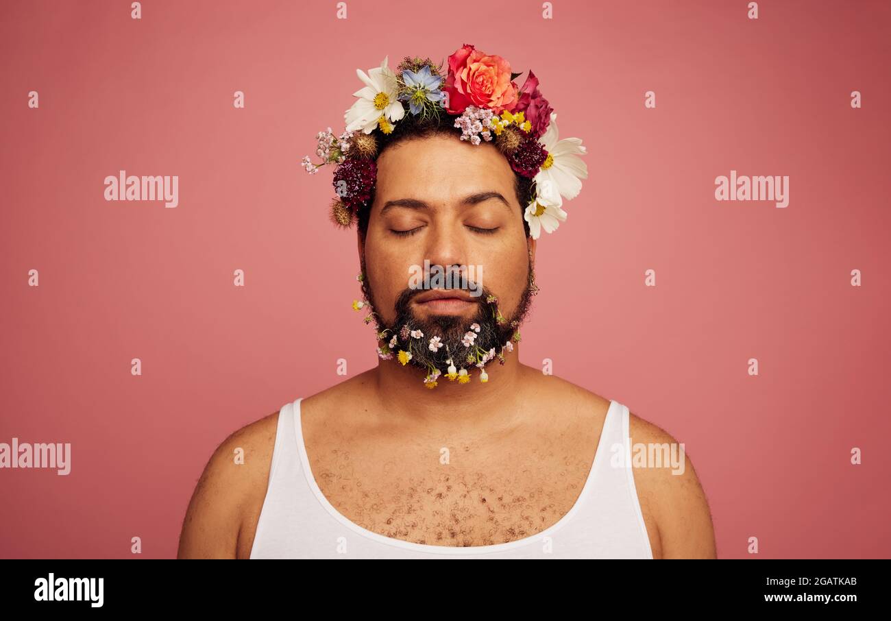 Uomo gay con fiori sulla testa e sulla barba. Sesso fluido maschio con occhi chiusi su sfondo rosa. Foto Stock