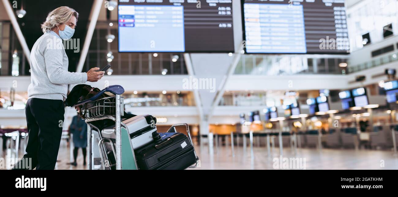 Uomo che indossa una maschera a piedi con il trolley bagagli al terminal dell'aeroporto. Viaggiatore maschile che usa il telefono cellulare all'aeroporto. Foto Stock