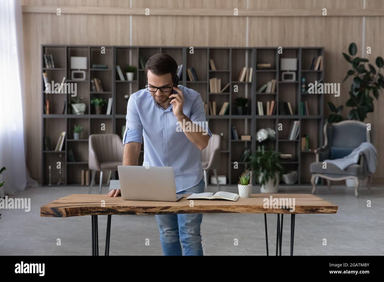 Un giovane uomo d'affari si trova in piedi da un computer portatile sul posto di lavoro, parla al telefono Foto Stock