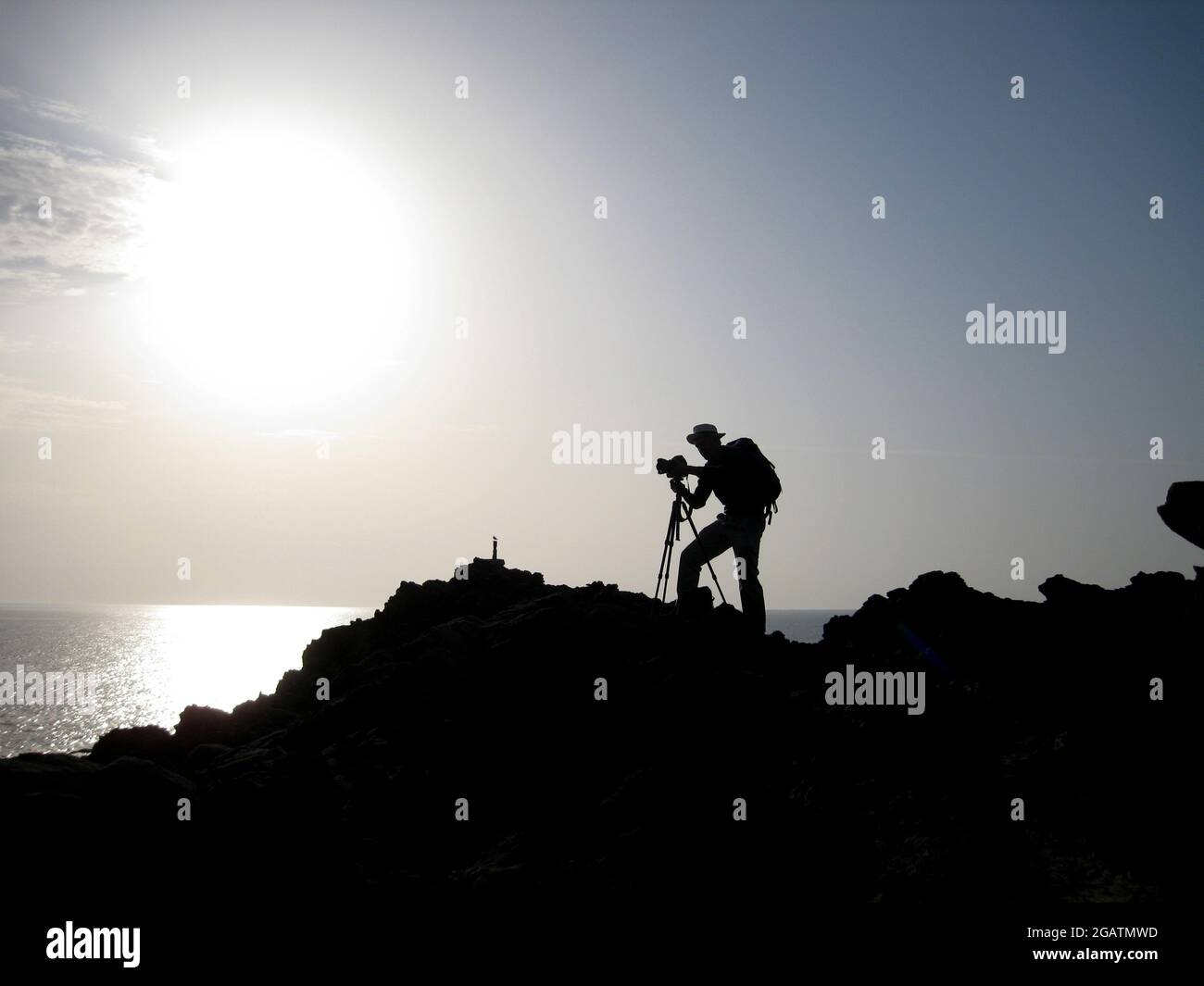 Il fotografo maschile si erge sulle rocce di Favàritx, Minorca, Spagna. Scatta fotografie con una macchina fotografica su un treppiede. Silhouette con mare in lontananza Foto Stock