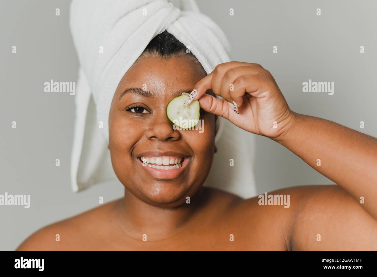 Ritratto di una giovane donna nera felice con asciugamano che si prende cura della sua pelle con un cetriolo naturale. Foto Stock