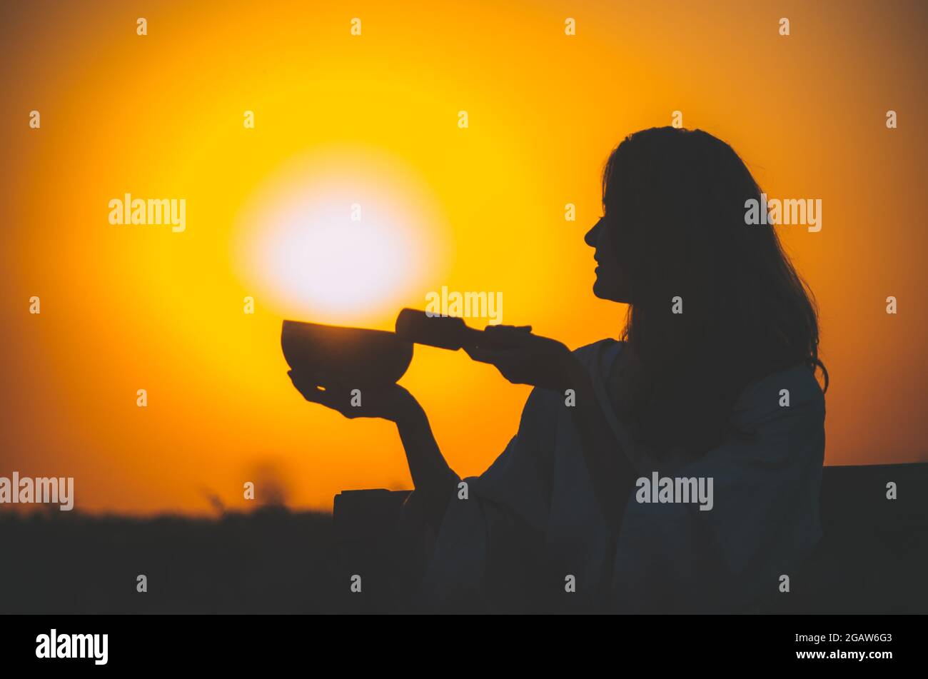 Silhouette cinematografica di donna che gioca una ciotola tibetana al meraviglioso tramonto arancione con disco solare. Concetto: Pace, tranquillità, zen, sano guarigione Foto Stock