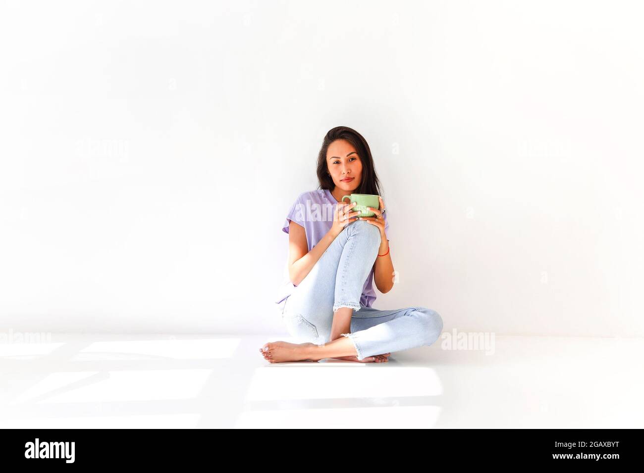 Donna etnica spensierata in abbigliamento domestico seduto sul pavimento in camera e gustare tè fresco durante la prima colazione a casa Foto Stock
