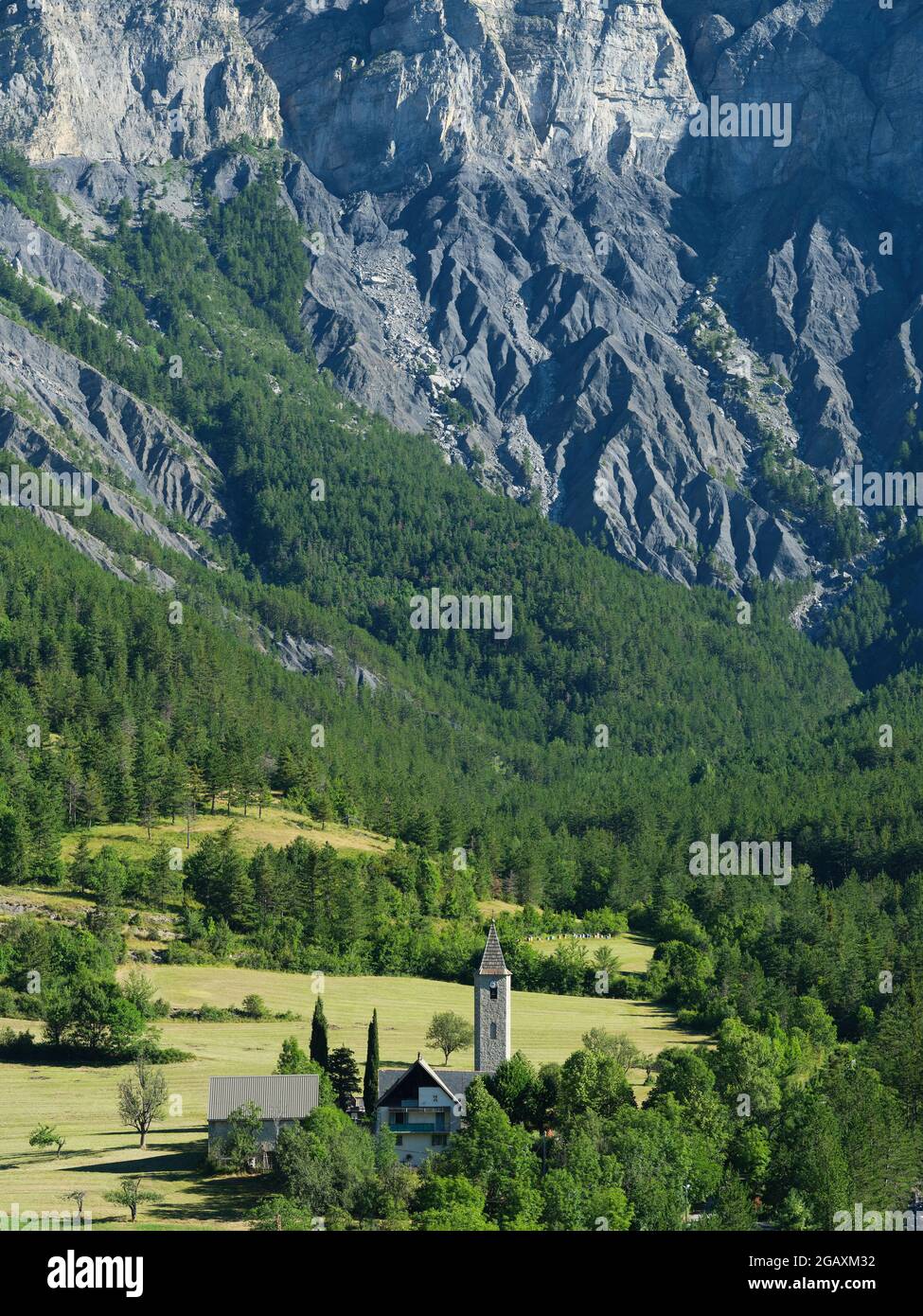 VISTA AEREA. Chiesa di Villeneuve d'Entraunes in piedi di una grande parete rocciosa. Alta Valle del Varo, Alpi Marittime, Francia. Foto Stock