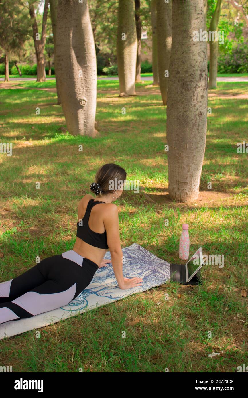 Immagine verticale di una giovane ragazza che fa la posa della cobra mentre si rilassa all'aperto nel parco cittadino in una giornata di sole in estate. Pilates Outdoor 2021. Foto Stock