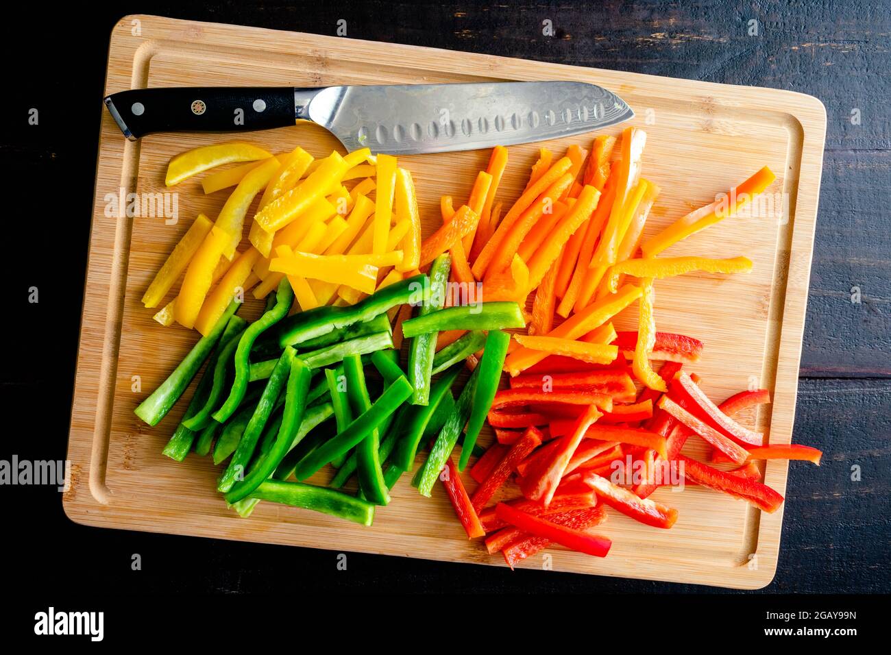 Peperoni rossi, arancioni, gialli e verdi tagliano in strisce sottili: Peperoni a campana tagliati in batonnets su un tagliere di bambù con un coltello dello chef Foto Stock