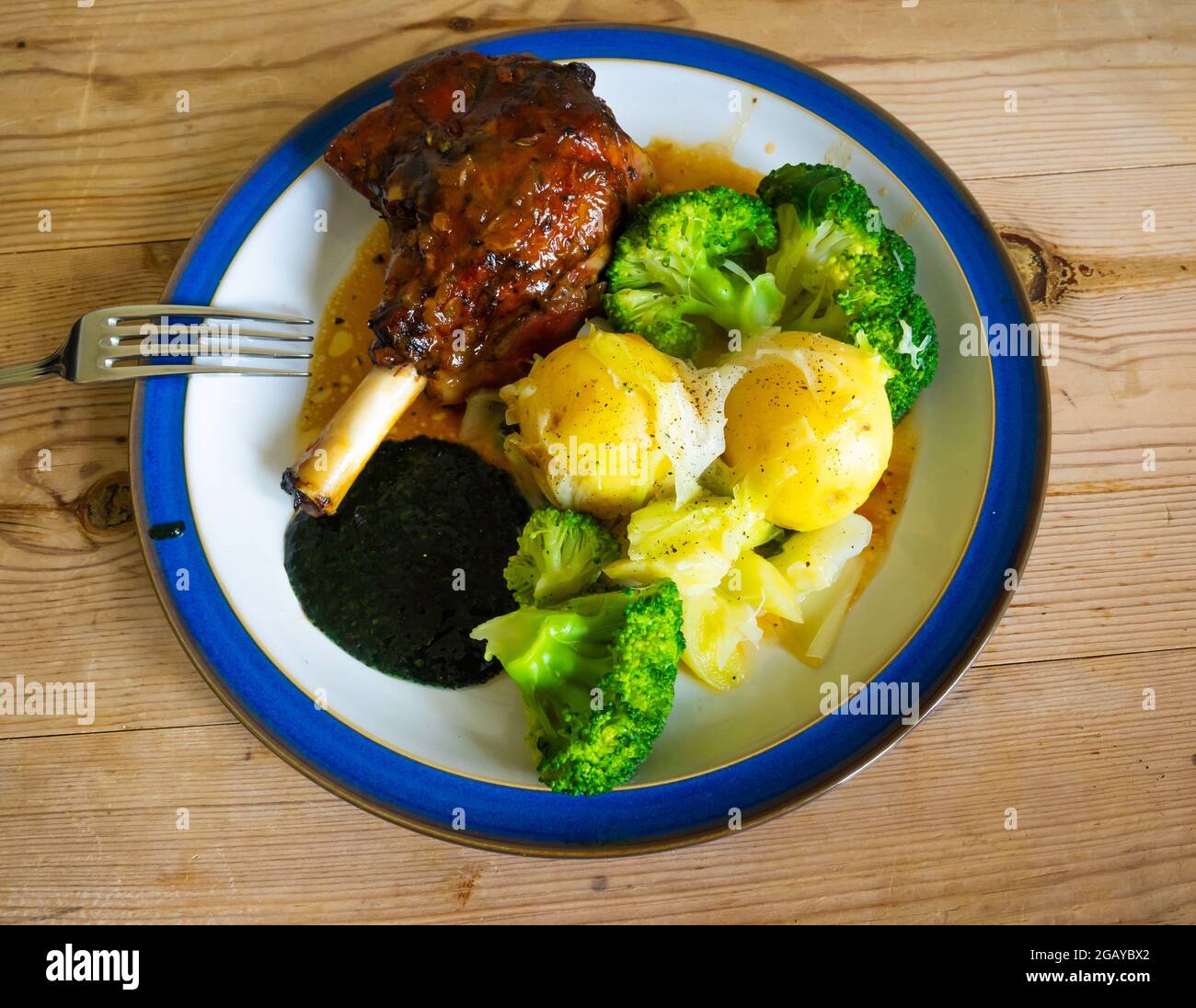 Pranzo inglese cena gamba di agnello bollito di patate novelle fagioli verdi broccoli e sugo su piatto bianco bordato blu su un tavolo di legno Foto Stock