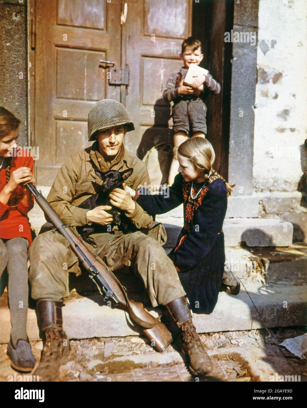 Seconda guerra mondiale seconda guerra mondiale colore soldato americano con bambini e un cane che fa amici seduti davanti a un gradino di una casa Foto Stock