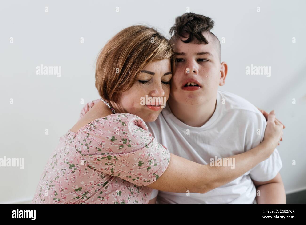 Madre abbracciando disabile ragazzo 17 anni in sedia a rotelle con paralisi cerebrale su sfondo bianco. Donna assiste la persona con bisogni speciali. Emotivo Foto Stock