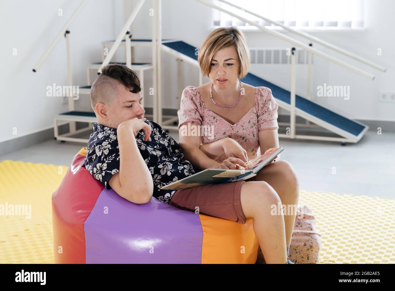 Bambini con disabilità leggere libro e comunicare. Ragazzo paralisi cerebrale che parla con la donna racconto terapeuta. Centro di riabilitazione, madre con Foto Stock