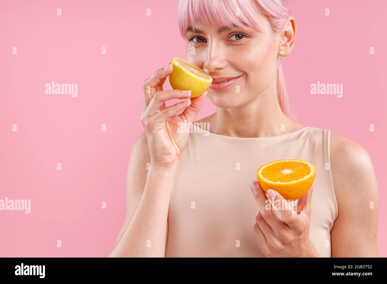 Ritratto di donna sorridente con capelli rosa che tiene metà dell'arancio maturo e odore di limone fresco, in posa isolata su sfondo rosa Foto Stock