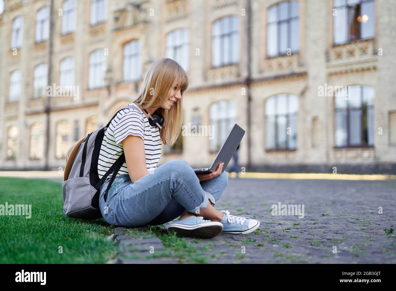 Bella giovane ragazza bionda scuola o studente universitario con zaino che indossa jeans utilizzando il computer portatile seduto su erba verde nel campus universitario. Ricerca Internet, comunicazione, concetto di tecnologia moderna Foto Stock