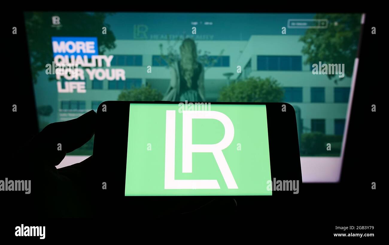 Persona che tiene il telefono mobile con il logo della società di cosmetici LR Health and Beauty Systems GmbH sullo schermo davanti alla pagina web. Mettere a fuoco il display del telefono. Foto Stock