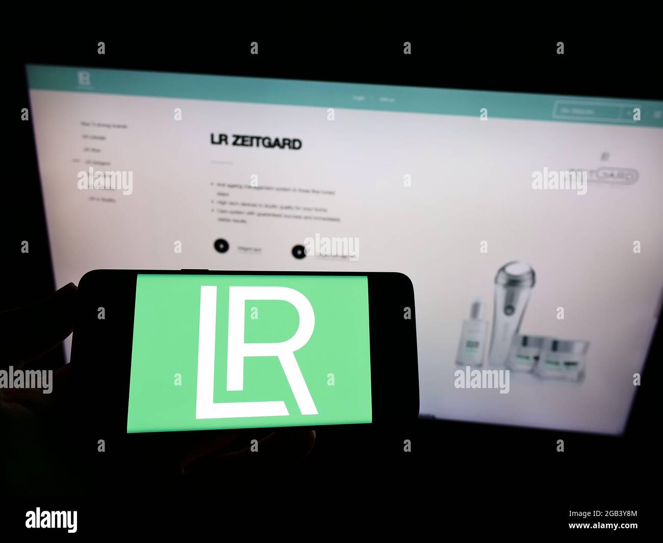 Persona che tiene il cellulare con il logo della società di cosmetici LR Health and Beauty Systems GmbH sullo schermo di fronte alla pagina web. Mettere a fuoco il display del telefono. Foto Stock