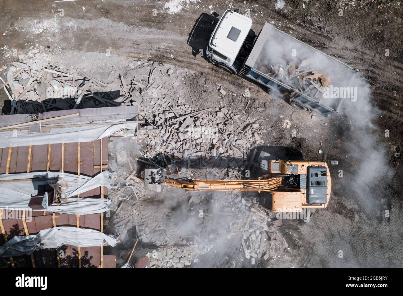 Demolizione di edifici da parte di escavatori industriali. Il secchio rompe i muri e il tetto della vecchia casa piena di polvere, vista aerea. Foto Stock