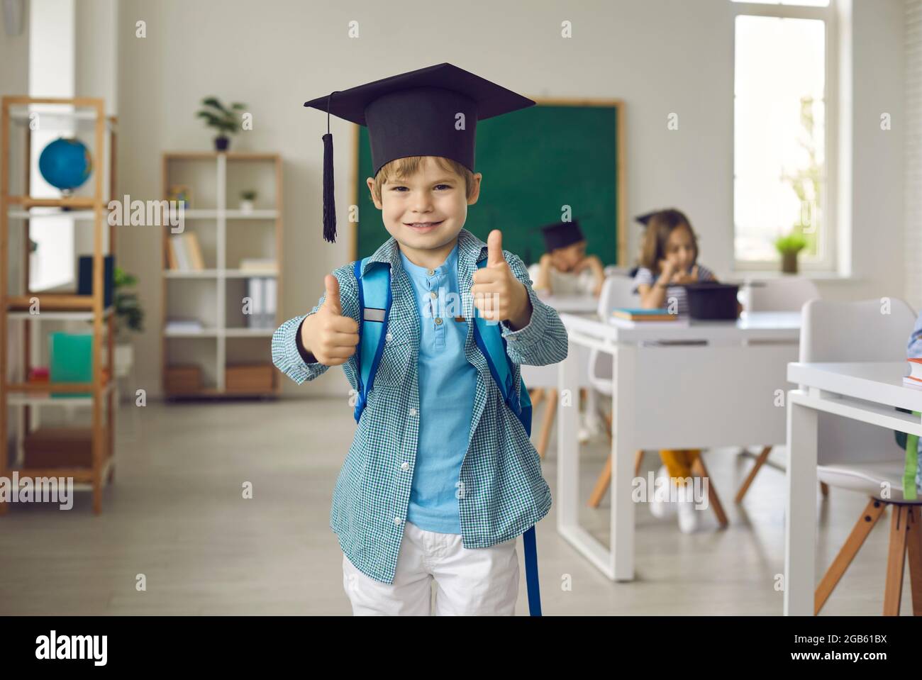 Felice studente di scuola elementare in un cappello accademico sorridente e dando un pollice in su Foto Stock