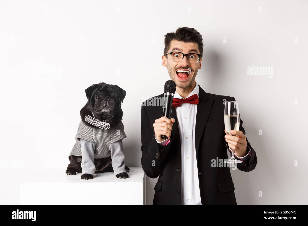 Felice giovane uomo dando un discorso e alzando champagne di vetro, in piedi con carino cane nero in costume da festa su sfondo bianco Foto Stock