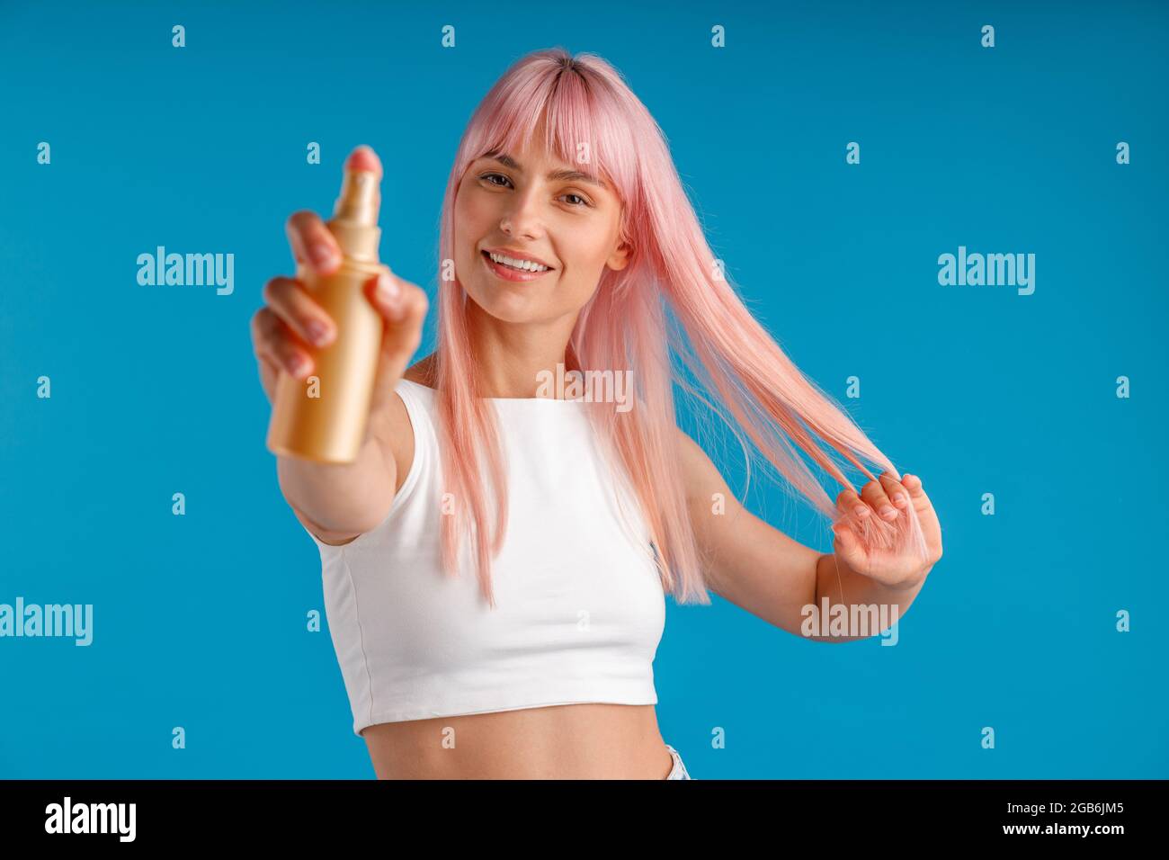 Ritratto di giovane donna con i capelli rosa sorridenti e mostrando una bottiglia di spray idratante alla macchina fotografica mentre si trova isolato su sfondo blu studio Foto Stock