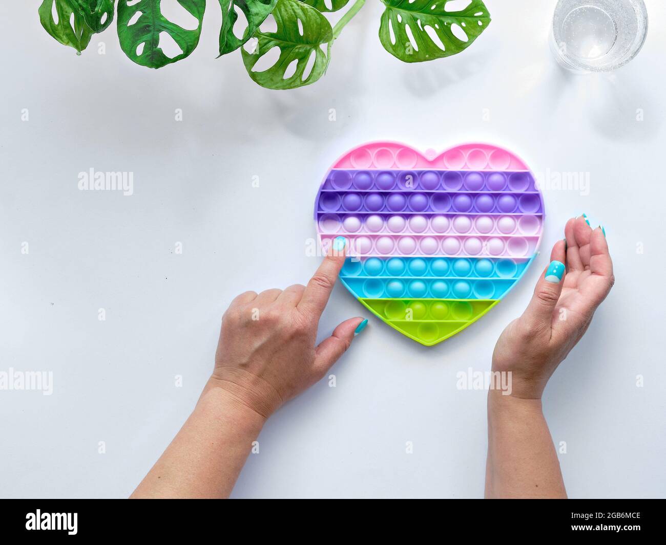 Giocattolo antistress a forma di cuore del papa in mani femminili. Fidget a concavità sensoriale semplice nei colori di LGBT. Piatto su sfondo bianco con motivi floreali Foto Stock