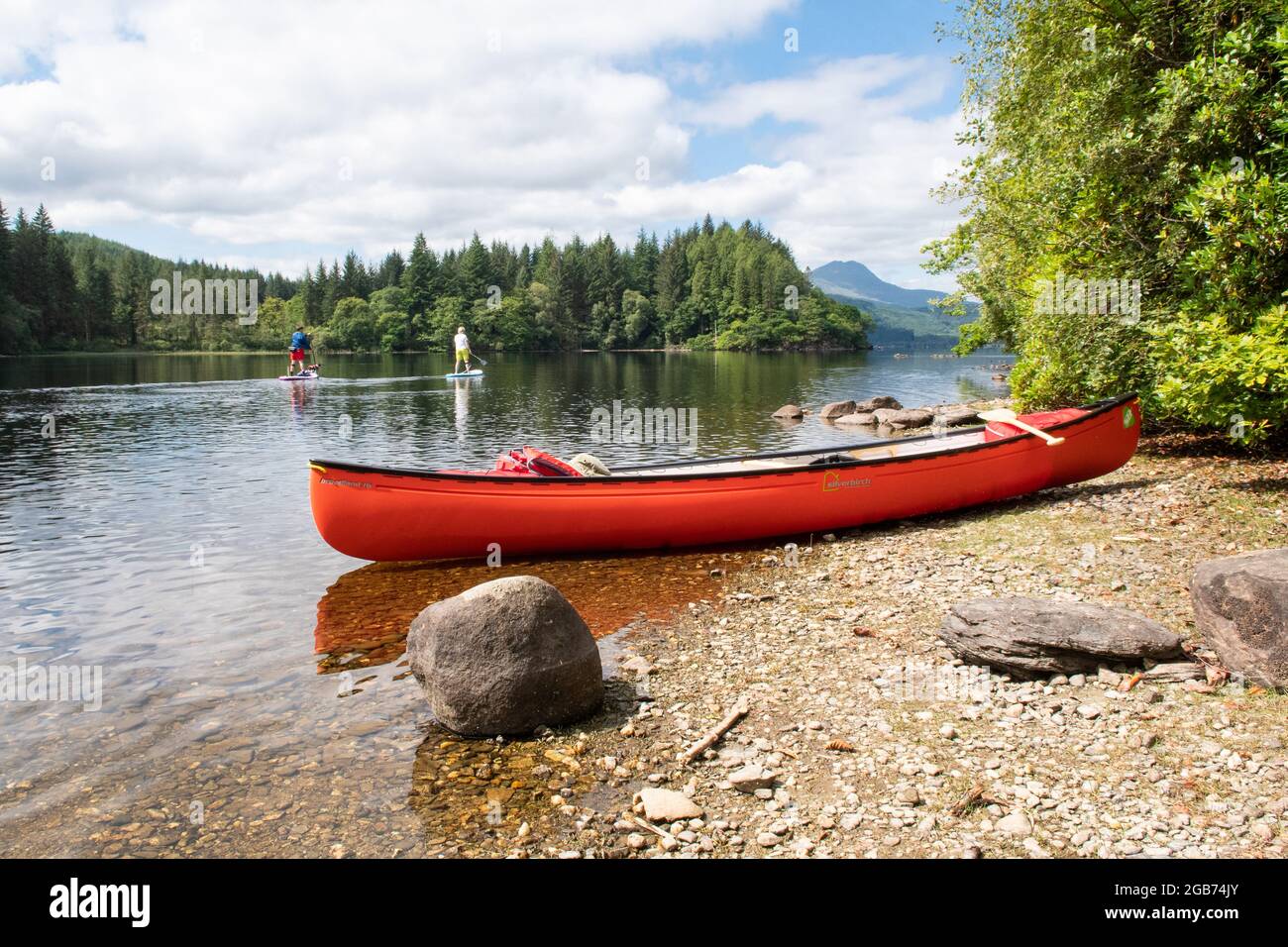 Canoa in Scozia - canoa arancione sulla piccola spiaggia e due paddle boarder passando con ben Lomond in lontananza, Loch Ard, Trossachs, Scozia, Foto Stock
