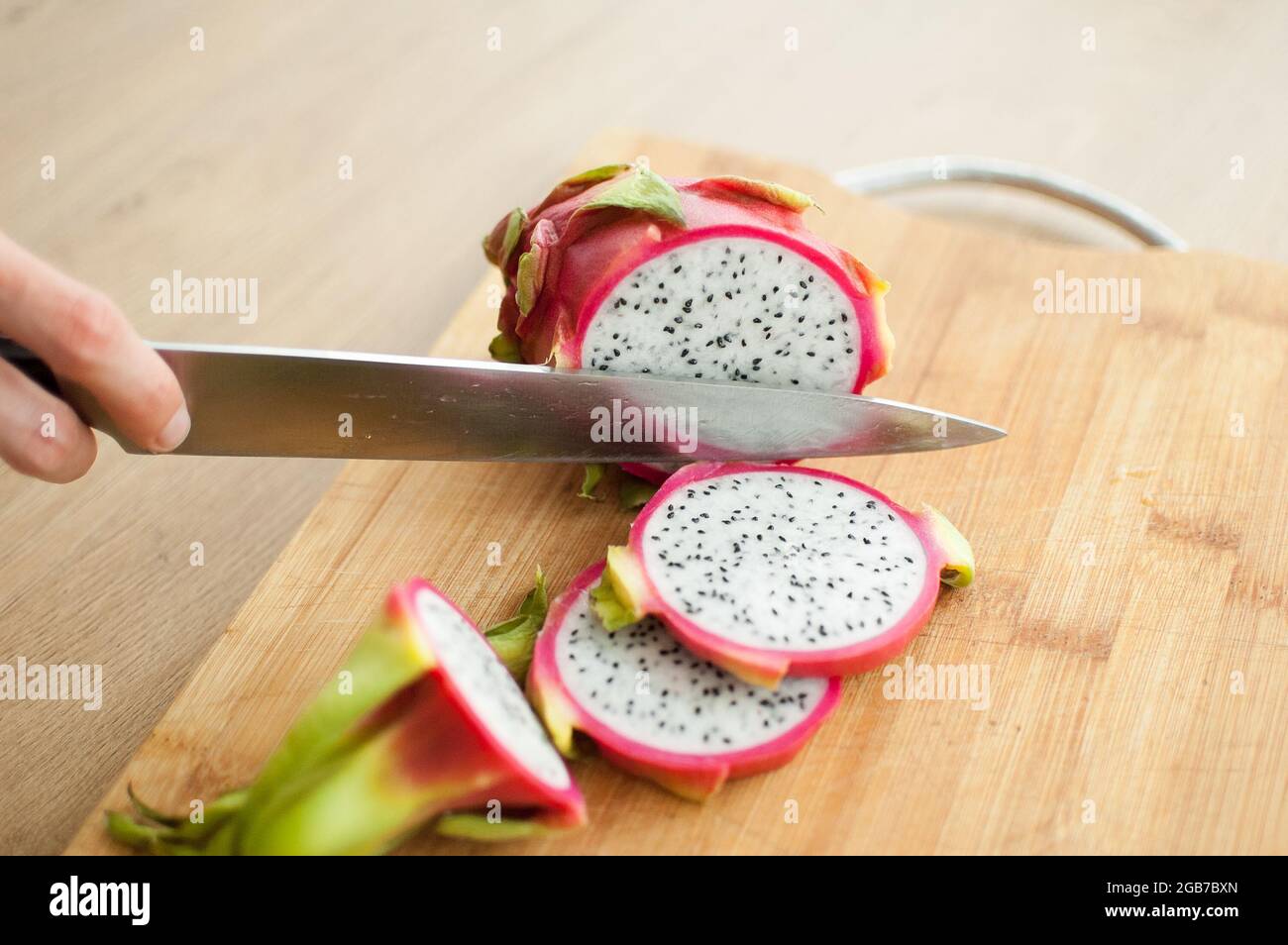 Le mani femminili sta tagliando un frutto di drago o pitaya con pelle rosa e polpa bianca con semi neri su bordo di legno tagliato sul tavolo. Frutta esotica Foto Stock