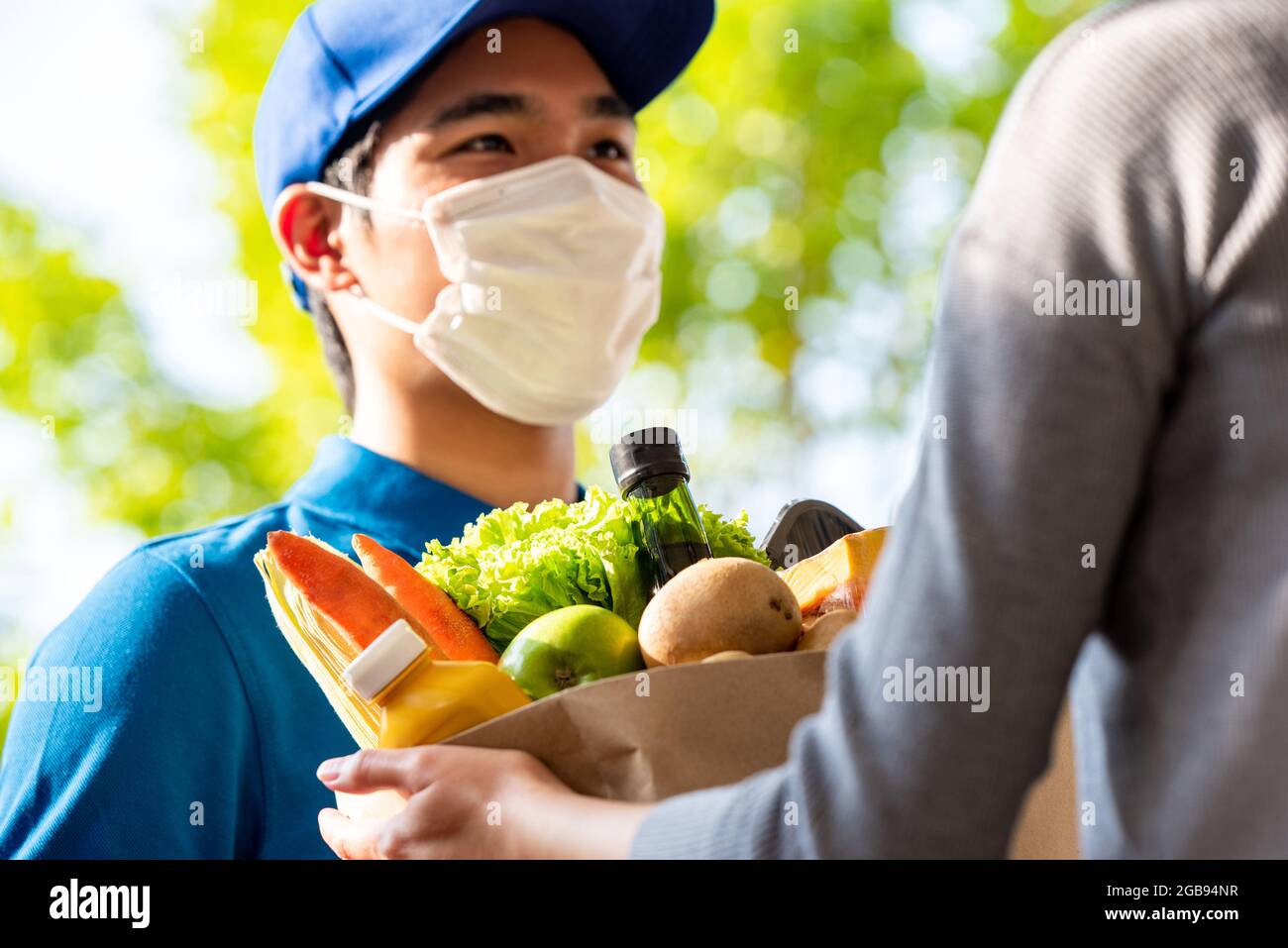 Uomo di deliveryman asiatico igienico che indossa la maschera facciale mentre consegna i generi alimentari al cliente a casa, consegna di cibo nel momento del concetto pandemico Foto Stock
