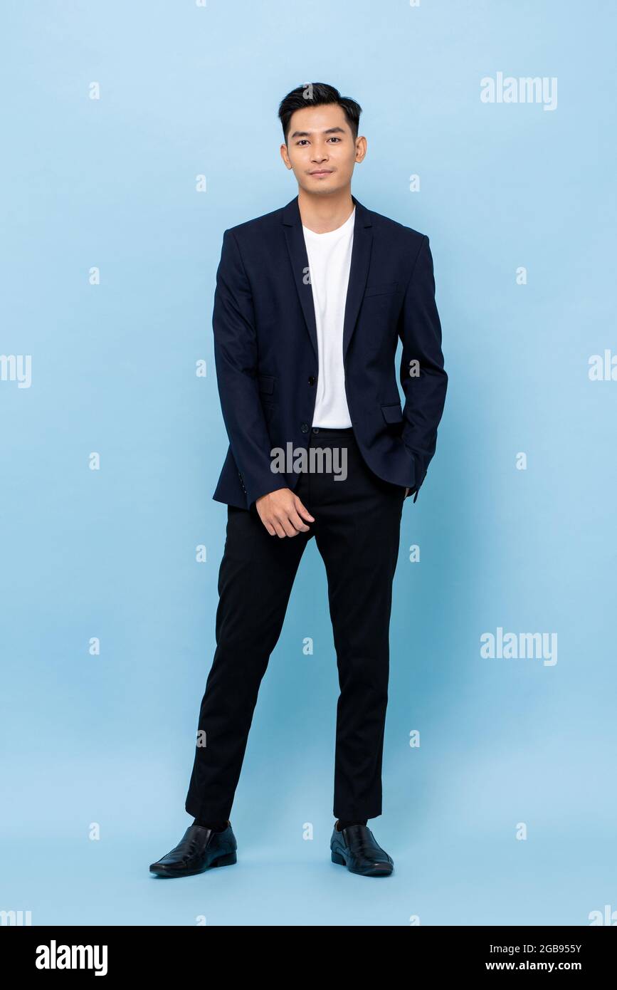 Ritratto a lunghezza intera di sorridente giovane bell'uomo asiatico in tuta semi formale in piedi con una mano in tasca su sfondo studio azzurro Foto Stock