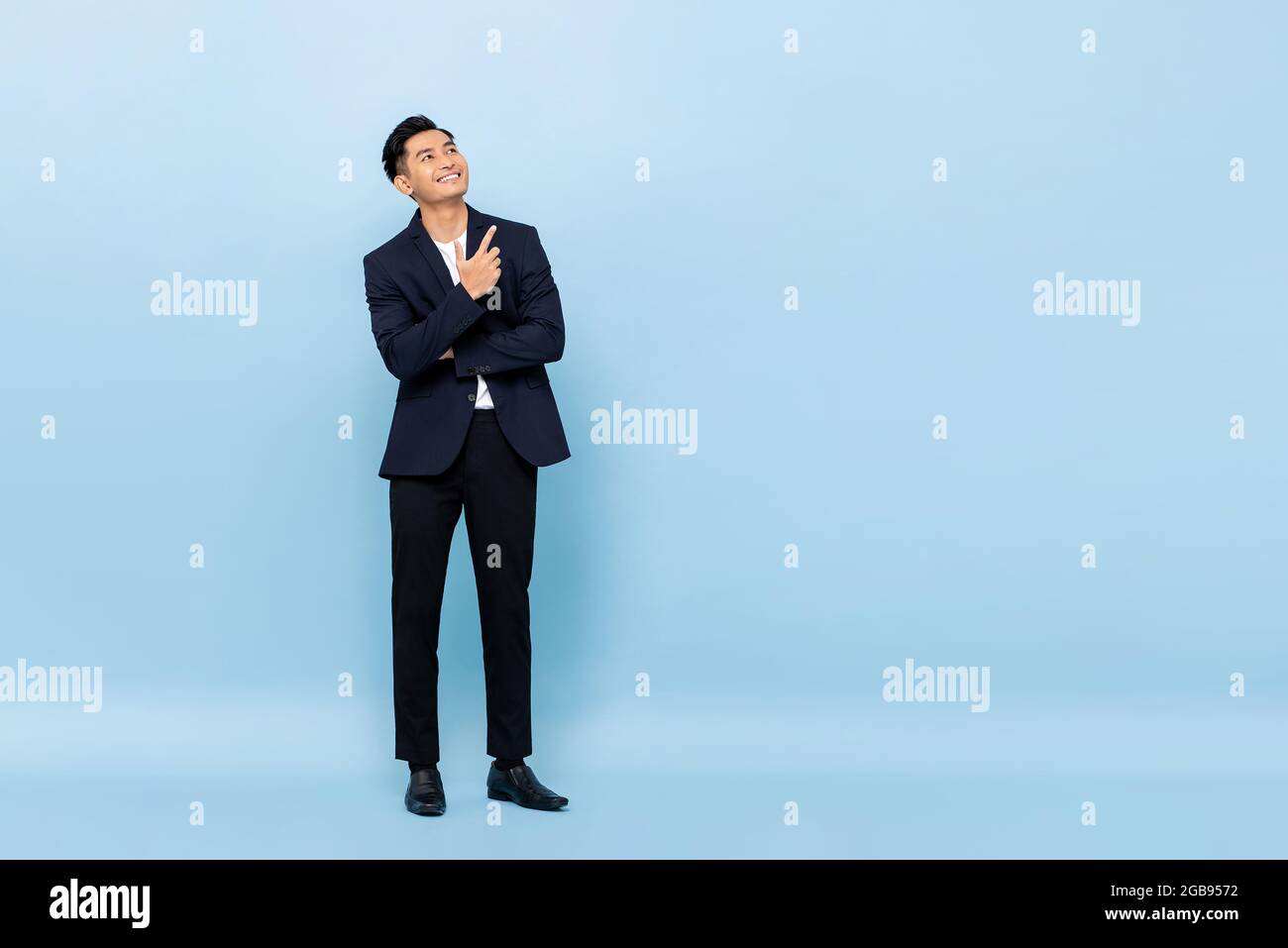 Ritratto a lunghezza intera di un giovane bell'uomo d'affari del sud-est asiatico che guarda in su e punta a copiare spazio su sfondo studio azzurro Foto Stock