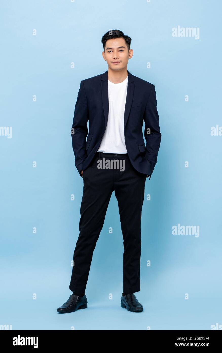 Ritratto a lunghezza intera di giovane bell'uomo d'affari asiatico in piedi e in posa con le mani in tasche su sfondo studio azzurro Foto Stock