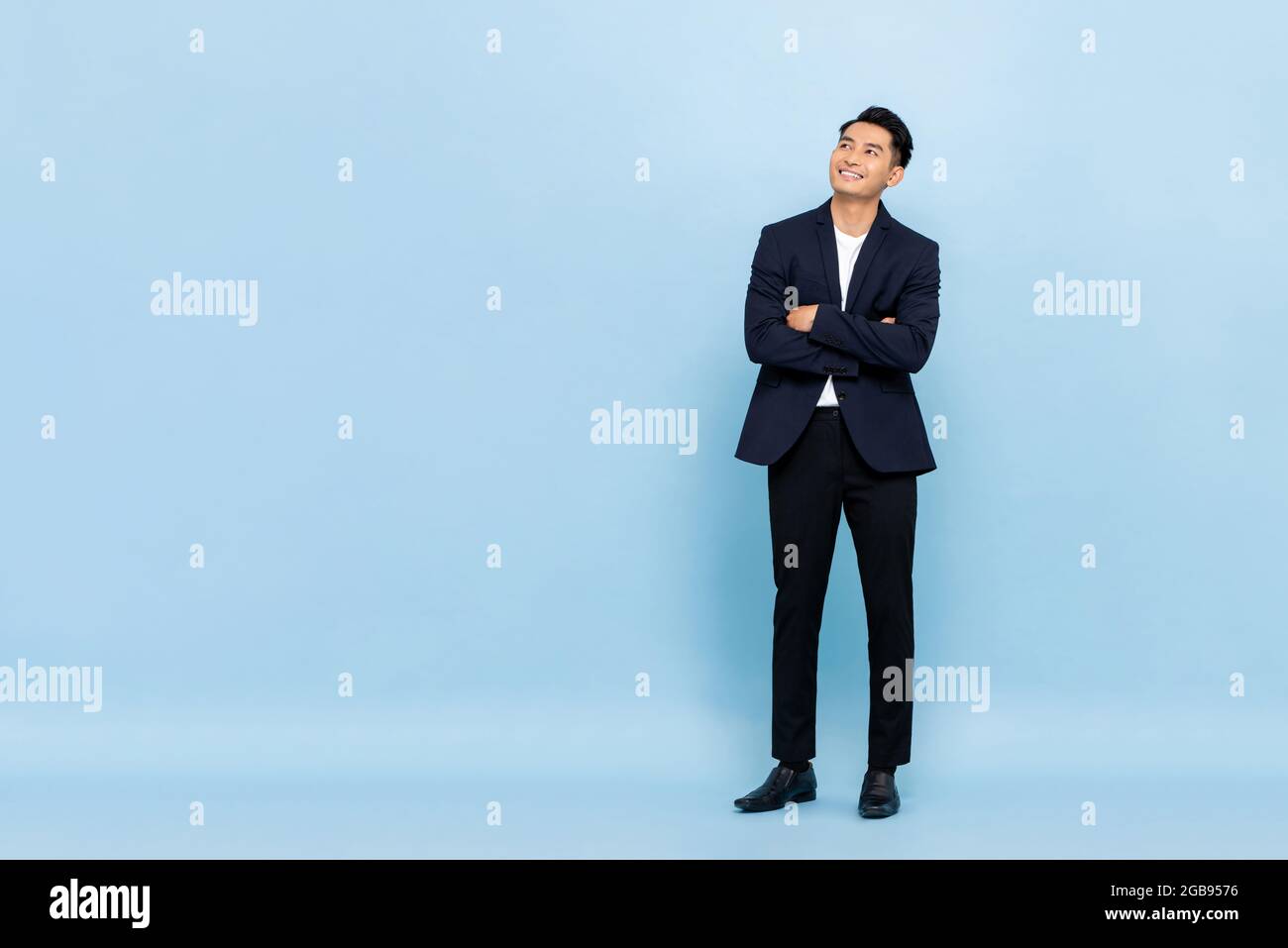 Ritratto a lunghezza intera di giovane bel uomo d'affari del sud-est asiatico con le braccia incrociate guardando fino a copiare spazio su sfondo studio azzurro Foto Stock