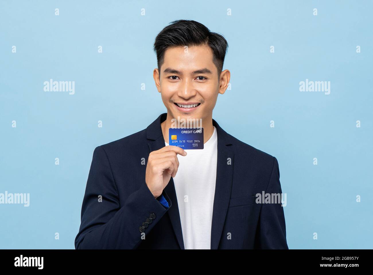 Giovane bell'uomo asiatico sorridente in blazer semi formale che tiene la carta di credito in blu chiaro isolato sfondo studio Foto Stock
